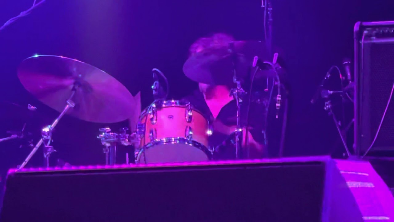 Drummer playing a drum kit on stage with purple stage lights.