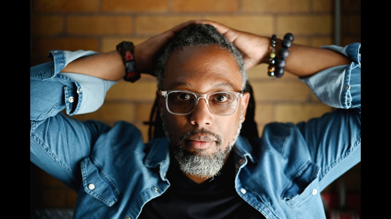 Man wearing glasses with arms behind head, seated indoors near brick wall