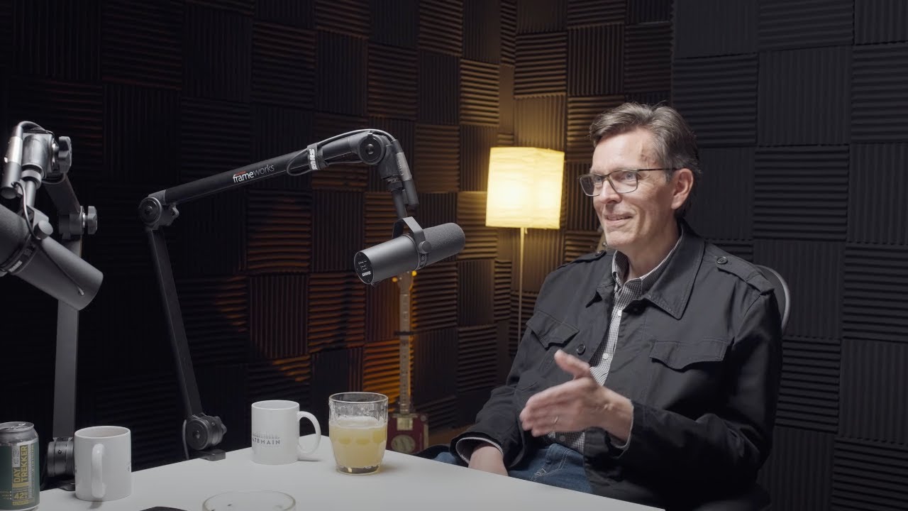 Man wearing glasses speaks beside studio microphones in a soundproof room.