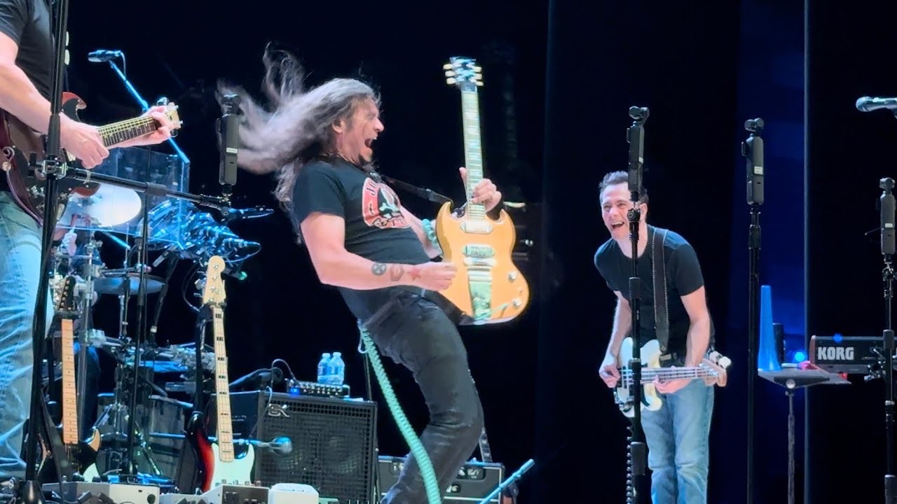 Guitarists play on a dark stage with microphones, drums, and amplifiers in view.